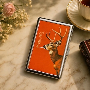May include: A rectangular metal cigarette case featuring a vibrant orange background with a graphic illustration of a deer with antlers smoking a cigarette. The case has a polished silver-toned frame and a hinged closure on the side.