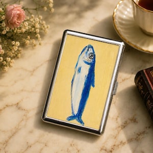 May include: A rectangular metal cigarette case featuring a painted illustration of a blue fish on a light yellow background. The case has a polished silver-tone frame and a hinged closure mechanism on the side, presenting a clean and functional design.