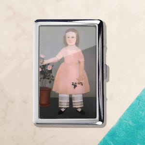 May include: A silver cigarette case featuring a vintage portrait of a young girl in a pink dress and white pants. The girl is holding a sprig of flowers and standing next to a potted rose bush. The case has a shiny, metallic finish.