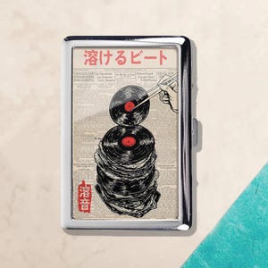 Pode incluir: Um porta-cigarros prateado com um gráfico de discos de vinil com rótulos vermelhos, empilhados uns sobre os outros. O design inclui texto japonês e uma mão usando pauzinhos para segurar um disco. O fundo é uma impressão de jornal.