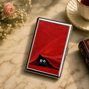 May include: A rectangular silver metal case featuring a vibrant red painted surface. The artwork depicts a small black cat with wide white eyes peeking out from underneath a textured, deep red fabric or blanket, creating a bold contrast.
