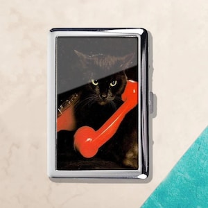May include: A silver metal cigarette case featuring a black cat holding a red telephone receiver. The cat has yellow eyes and is set against a dark background. The case has a rectangular shape.