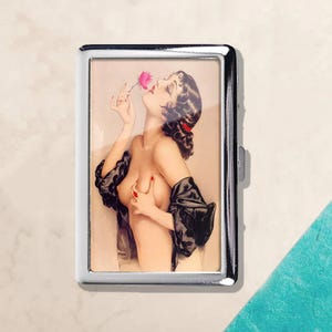May include: A silver cigarette case featuring a vintage pin-up illustration. The artwork depicts a woman with dark hair and red lipstick holding a pink flower. She is wearing a black robe and has red nail polish.