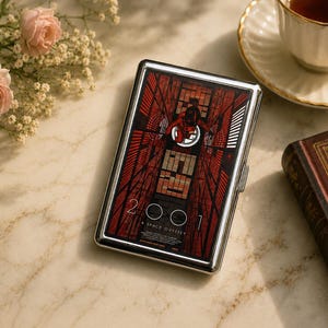 May include: A rectangular silver metal cigarette case featuring a graphic print of the 2001: A Space Odyssey movie poster. The design displays a red and black geometric corridor with an astronaut figure and the text 2001 A SPACE ODYSSEY.
