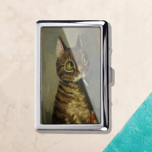 May include: A silver metal cigarette case featuring a whimsical illustration of a tabby cat with large green eyes smoking a cigarette. The cat is brown and tan with a red lighter in the image.