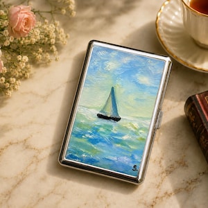 May include: A rectangular silver metal cigarette case featuring an oil painting on the front. The artwork depicts a sailboat on a textured blue and green sea under a bright sky with visible brushstrokes. The letter S is signed in the bottom corner.