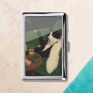 Puede incluir: Estuche de cigarrillos plateado con una ilustración de un gato blanco y negro con collar de perlas fumando un cigarrillo, sentado en un sofá verde. Una pequeña mesa con bebida y aperitivos.
