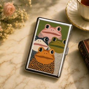 May include: A rectangular metal case with a polished silver frame featuring a graphic illustration of four frogs in shades of pink, blue, green, and orange with black spots. The frogs are stacked against a solid green background.