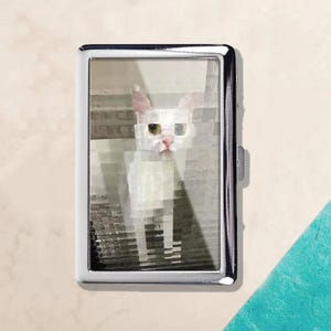 May include: A silver metal cigarette case with a pixelated image of a white cat with pink ears and yellow eyes. The cat is centered within the case, which has a rectangular shape and a hinged closure.
