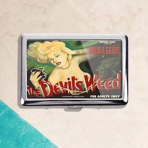 Puede incluir: Un estuche de cigarrillos de metal plateado con un cartel de película vintage de "The Devil's Weed". El cartel presenta a una mujer rubia, el texto "Lola Leeds" y la frase "For Adults Only".