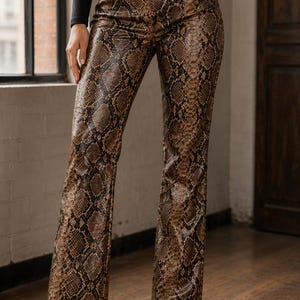 Puede incluir: Pantalones de talle alto con estampado de piel de serpiente en marrón y negro. Estos pantalones tienen una silueta de pierna acampanada, cierre de botón frontal, trabillas para cinturón y bolsillos laterales. Acabado liso y texturizado.