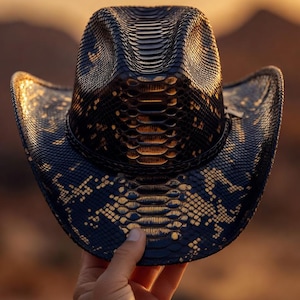 Pode incluir: Um chapéu de cowboy preto com um padrão de textura de pele de cobra em toda a superfície. O chapéu tem uma copa estruturada com um vinco central e uma aba curva, refletindo luz dourada ao longo das bordas do material.