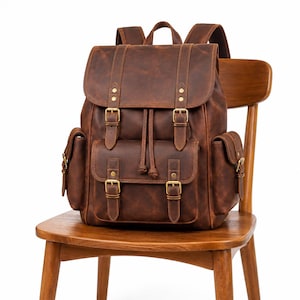 May include: A brown leather backpack featuring a top flap with two buckle straps, a drawstring closure, and a front pocket with two additional buckle straps. The bag includes two side pockets and brass-toned metal hardware throughout the design.