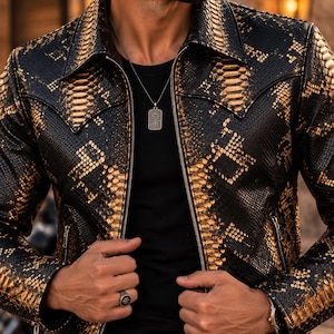 Puede incluir: Una chaqueta con estampado de piel de serpiente en negro y dorado, cuello en punta, cremallera frontal completa y bolsillos laterales con cremallera. La prenda se lleva sobre una camiseta negra lisa y se complementa con un collar de colgante rectangular.