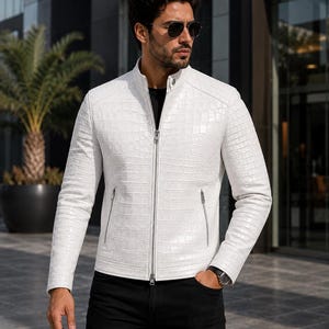 Könnte beinhalten: Eine weiße Lederjacke mit texturiertem Krokodilledermuster, ausgestattet mit einem Stehkragen, einem durchgehenden silbernen Frontreißverschluss und zwei seitlichen Reißverschlusstaschen. Die Jacke wird über einem schwarzen Oberteil getragen.