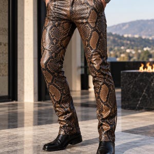 May include: Fitted trousers featuring a brown and black snakeskin pattern with a glossy finish. The trousers include a standard waistband with belt loops, a front button closure, and straight legs, paired with black leather boots.