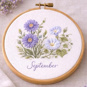 以下が含まれることがあります： 木製のフレームに刺繍された花の芸術作品。紫と青の花と緑の葉が特徴です。「September」の文字が刺繍されています。刺繍は木製のフレームに収められています。