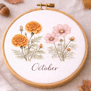 Può includere: Ricamo floreale in un telaio di legno, con calendule arancioni e cosmos rosa. La parola "October" è ricamata sotto i fiori. Foglie autunnali e una ghianda sullo sfondo.