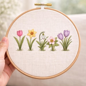 May include: Embroidered floral hoop art featuring a row of spring flowers: a pink tulip, yellow daffodil, white snowdrop, pink flower, and purple crocus. The flowers are stitched on a cream-colored fabric, set in a wooden hoop.