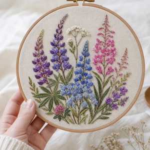 May include: A hand-embroidered floral design in a wooden hoop. The artwork showcases a selection of colourful flowers, such as purple lupins, blue forget-me-nots, and pink fireweed, with green leaves. The embroidery is on a cream-coloured fabric.