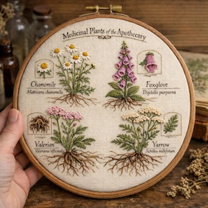 May include: Embroidered hoop art featuring four medicinal plants: chamomile, foxglove, valerian, and yarrow. Each plant is labeled with its common and scientific name. The artwork is set against a beige fabric background within a wooden hoop.