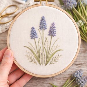 Pode incluir: Bordado floral em um bastidor de madeira. O design apresenta três flores de jacinto-uva azuis com caules e folhas verdes. O bordado é em tecido creme, com um fecho de latão.