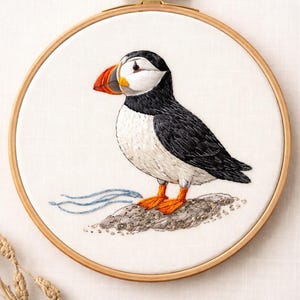 以下が含まれることがあります： 木製のフレームに入った、刺繍されたツノメドリのアートワーク。鳥は白と黒の体、オレンジ色の足、大きくカラフルなくちばしを持っています。背景は明るい生地で、フレームは真鍮製の留め金で固定されています。
