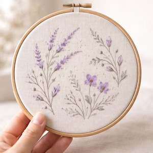 May include: A circular embroidery hoop with a floral design. The design features lavender and other purple flowers with green leaves, stitched on a cream fabric. The wooden hoop is held by a hand.