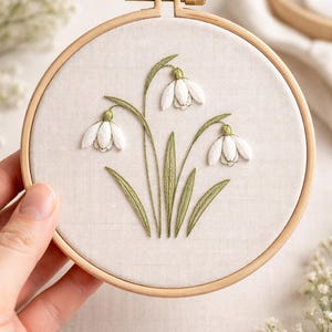 Op de afbeelding: Een handgeborduurde borduurring met een ontwerp van witte sneeuwklokjes met groene stelen en bladeren. De borduursel is geplaatst tegen een beige stoffen achtergrond, omlijst door een houten ring.