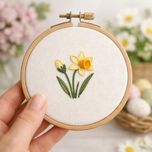 May include: A hand-stitched embroidery hoop featuring a yellow daffodil flower and bud. The flower has a yellow centre and petals, with green leaves and stem. The embroidery is set in a wooden hoop, creating a charming, handcrafted piece.