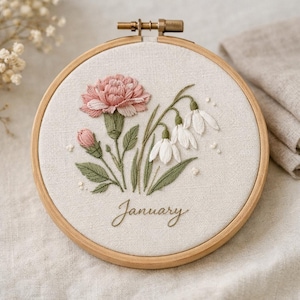 Op de afbeelding: Geborduurde bloemenkunst in een borduurring met een roze anjer, witte sneeuwklokjes en groene bladeren. Het woord "January" is eronder geborduurd. De borduursel zit in een houten ring.