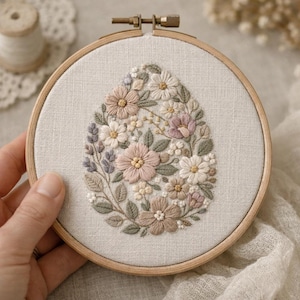 May include: An embroidered floral design in a wooden hoop. The design is an egg shape filled with flowers in pink, cream, and green. The embroidery is on a light beige fabric.