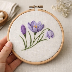 May include: Hand-embroidered floral art in a wooden hoop. The design features three purple crocus flowers with green stems and leaves, stitched on a cream-colored fabric. The embroidery hoop is approximately 6 inches in diameter, perfect for wall decor.