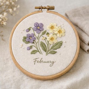 Puede incluir: Un bastidor de bordado floral con un ramo de flores moradas y amarillas con hojas verdes. La palabra "February" está bordada debajo de las flores. El bordado está en un bastidor de madera.