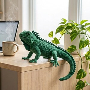 May include: A handcrafted, green crochet iguana sits on a wooden desk next to a laptop and a steaming mug. A potted plant with trailing vines is in the background. The iguana is detailed with textured scales and a spiky dorsal ridge.