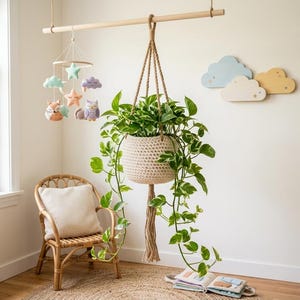 Pode incluir: Um suporte de plantas em crochê bege com uma planta jiboia verde e branca, suspenso por uma haste de madeira. Ao lado, um móbile com estrelas, nuvens, uma raposa e uma coruja de pelúcia. Três decorações de parede de madeira em forma de nuvem.