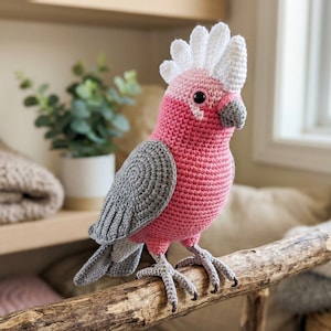 May include: A handmade crochet bird in pink, gray, and white, perched on a wooden branch. The bird has a distinctive crest and is likely a cockatoo. The craft item is displayed indoors.