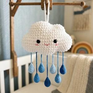 May include: A handmade white crochet cloud mobile with blue raindrop accents. The cloud has a smiling face with black eyes and pink cheeks. The mobile is suspended from a wooden frame, perfect for a nursery.
