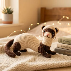 Puede incluir: Un juguete de hur&oacute;n tejido a ganchillo en crema y marr&oacute;n, con nariz rosa y bigotes negros, descansa sobre una manta texturizada. El animal de peluche hecho a mano est&aacute; cerca de una pila de libros, lo que sugiere un tema literario y acogedor. El hur&oacute;n mide unos 30 cm de largo.
