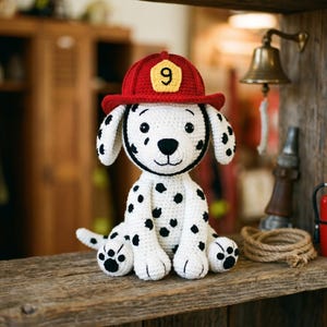 Pode incluir: Um cão dálmata de crochê com um chapéu de bombeiro vermelho com o número 9. O cão tem manchas pretas e está sentado numa prateleira de madeira. À direita, há um pequeno extintor vermelho.