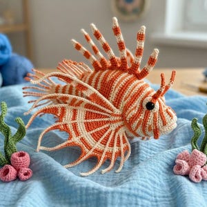 May include: A crocheted lionfish toy featuring orange and cream stripes, prominent spiky fins, and a black eye. The fish is accompanied by two small crocheted green seaweed stalks and clusters of pink coral, all resting on a light blue fabric.