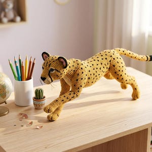 May include: A handmade, yellow crochet cheetah with black spots, posed in a running position. The cheetah is on a light wood table, with a globe, pencils, and a small cactus nearby. The cheetah's tail has a black tip.