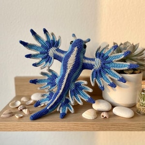 May include: A crocheted blue dragon sea slug plush toy featuring a central body with a light beige stripe and multiple radiating, pointed appendages in shades of dark blue, medium blue, and light blue with white tips and black eyes.
