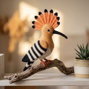 Puede incluir: Un pájaro de ganchillo hecho a mano con detalles en naranja, negro y blanco, posado en una rama de madera. El pájaro tiene un pico negro largo y curvado y una cresta naranja. También se ve una pequeña planta suculenta en una maceta de cerámica.