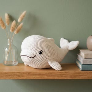 May include: A handmade, white crochet whale toy with black eyes and a smiling mouth. The whale is on a wooden shelf with a vase of dried flowers and a candle holder. The background is a muted green wall.