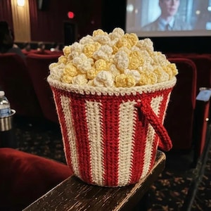 May include: A crocheted popcorn bucket featuring vertical red and cream stripes with a handle on the side. The bucket is filled with textured, three-dimensional crocheted popcorn kernels in shades of pale yellow and off-white, creating a realistic look.