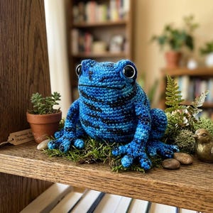 May include: A handmade blue and black crochet frog figurine with large black eyes. The frog sits on a bed of green moss, with a small potted succulent and a decorative bird nearby. The frog is a whimsical home decor item.