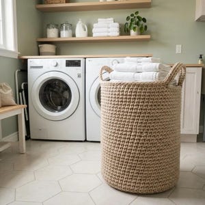 May include: A large, cylindrical laundry basket made of thick, beige woven rope with two integrated handles. The basket is filled with a stack of folded white towels, showcasing a textured, sturdy construction suitable for holding laundry items.