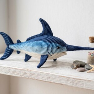 May include: A handmade crochet swordfish in shades of blue and white rests on a white shelf. The toy fish features a long, pointed snout and a dorsal fin. A small glass bottle with a cork stopper, filled with sand and shells, sits nearby.