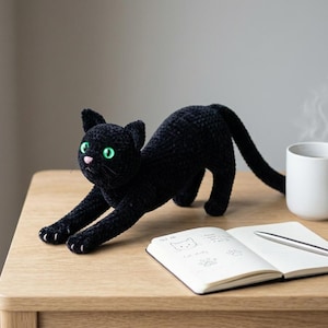 Puede incluir: Un gato negro de ganchillo con ojos verdes y nariz rosa se estira sobre una superficie de madera. Una taza blanca con vapor, una pequeña suculenta y un cuaderno abierto con un bolígrafo también están sobre la mesa. El gato es el punto focal.